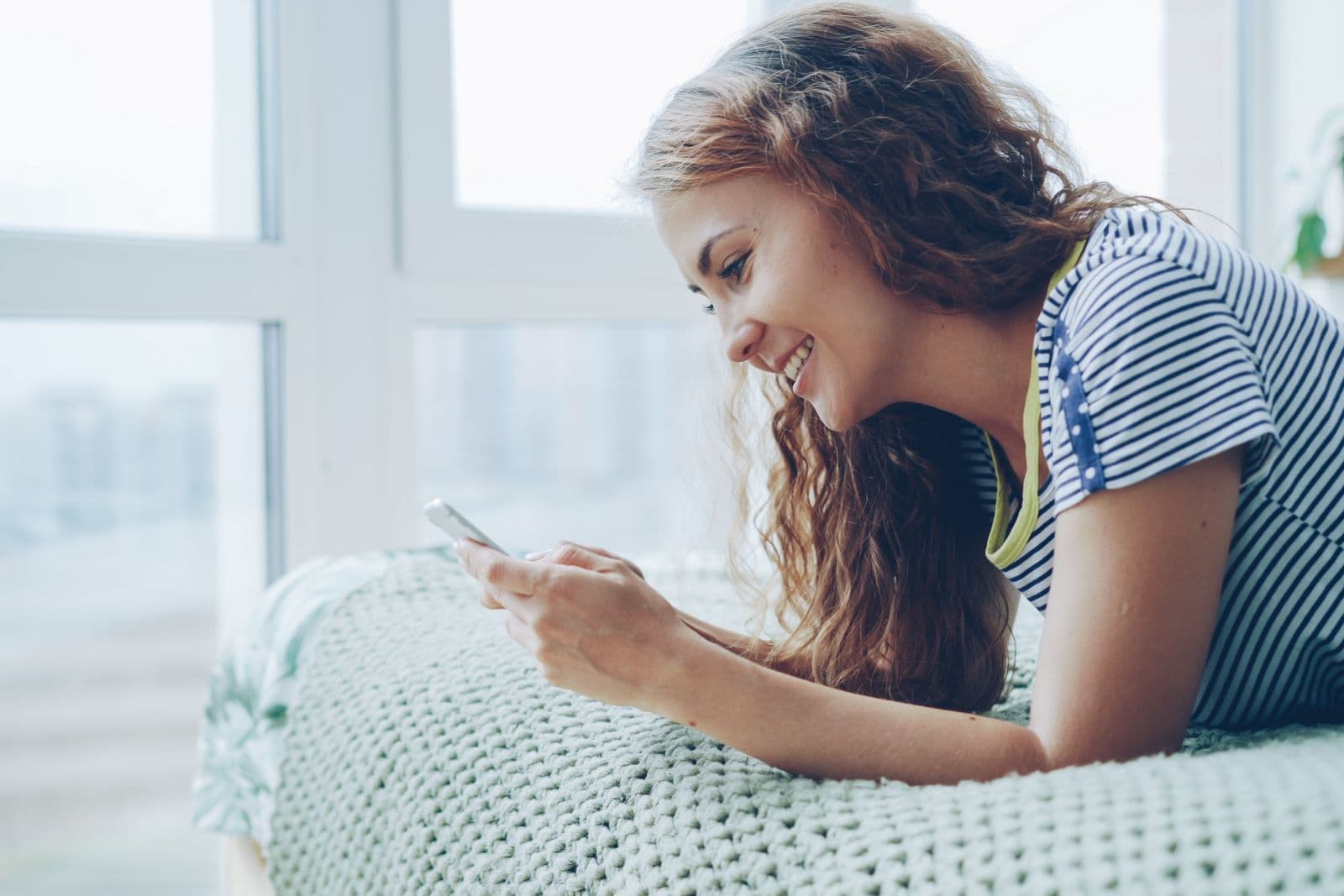 Mulher sorrindo enquanto conversa pelo celular, deitada na cama em casa