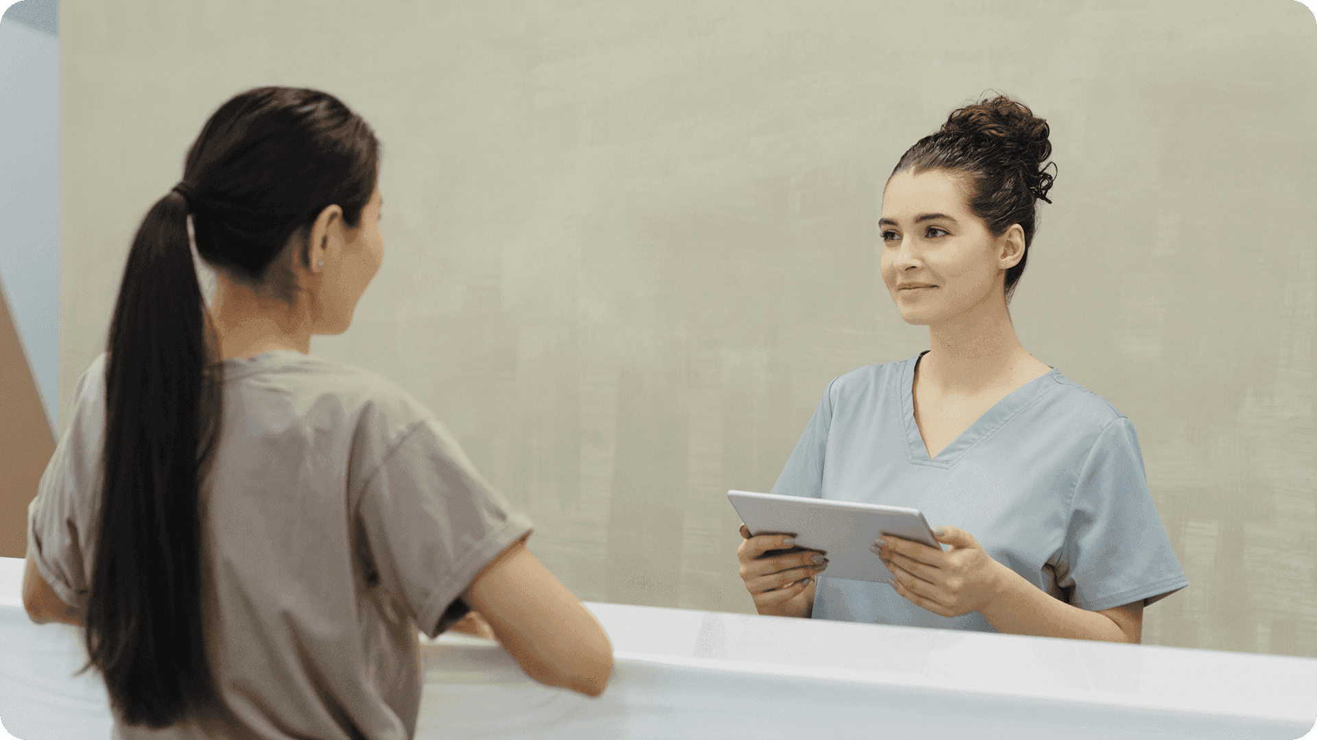 Recepcionista de scrubs com tablet atendendo paciente no balcão de uma clínica